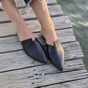 May include: Black leather slip-on mules with a pointed toe and crocodile texture. The shoes are on a wooden surface, with a blurred background. The mules are a stylish footwear option.