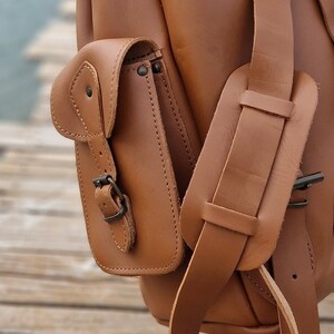 Puede incluir: Una mochila de cuero marr&oacute;n con una solapa grande y un bolsillo m&aacute;s peque&ntilde;o en la parte delantera. La mochila tiene una hebilla en el bolsillo delantero y una correa que se coloca sobre el hombro.