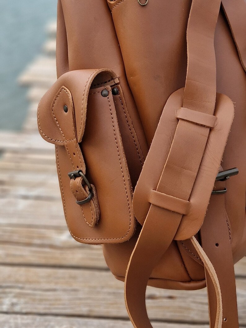 Puede incluir: Una mochila de cuero marr&oacute;n con una solapa grande y un bolsillo m&aacute;s peque&ntilde;o en la parte delantera. La mochila tiene una hebilla en el bolsillo delantero y una correa que se coloca sobre el hombro.