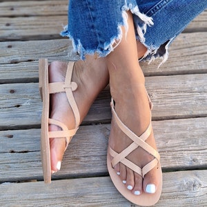 Op de afbeelding: Een paar lichtbruine leren sandalen met gekruiste banden. De sandalen worden gedragen door een vrouw.