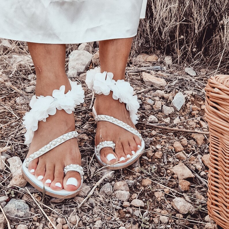 May include: White sandals with flower embellishments. The sandals have a white strap that goes around the ankle and a white strap that goes across the foot. The sandals are decorated with white flowers.