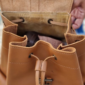Puede incluir: Una mochila de cuero marr&oacute;n con cierre de cord&oacute;n y hebilla. La mochila est&aacute; abierta y el interior est&aacute; forrado con un tejido marr&oacute;n claro.