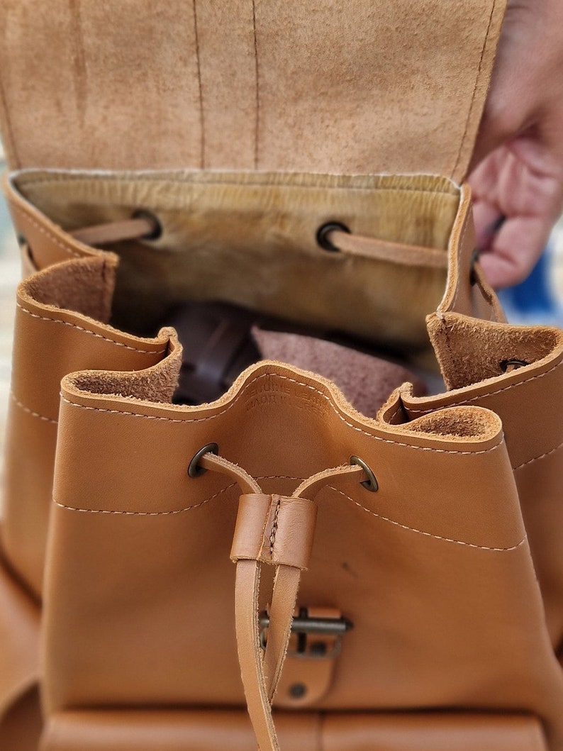 Puede incluir: Una mochila de cuero marr&oacute;n con cierre de cord&oacute;n y hebilla. La mochila est&aacute; abierta y el interior est&aacute; forrado con un tejido marr&oacute;n claro.