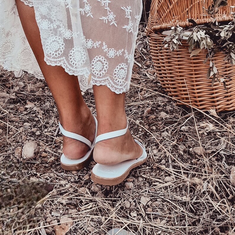 May include: A pair of white leather sandals with thin straps. The sandals are worn by a person wearing a white lace dress. The sandals are on a bed of dry grass.