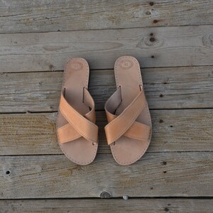 May include: A pair of tan leather sandals with criss-cross straps. The sandals are on a wooden surface.