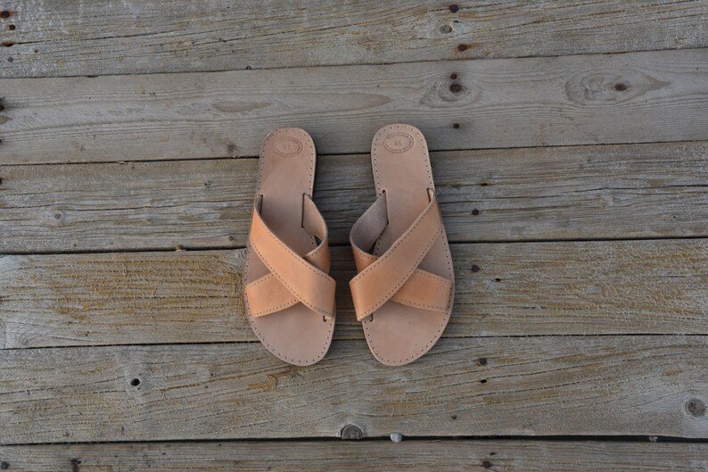 May include: A pair of tan leather sandals with criss-cross straps. The sandals are on a wooden surface.
