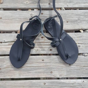 K&ouml;nnte beinhalten: Ein Paar schwarze Ledersandalen mit Riemen, die &uuml;ber Fu&szlig; und Kn&ouml;chel verlaufen. Die Sandalen befinden sich auf einer Holzoberfl&auml;che.