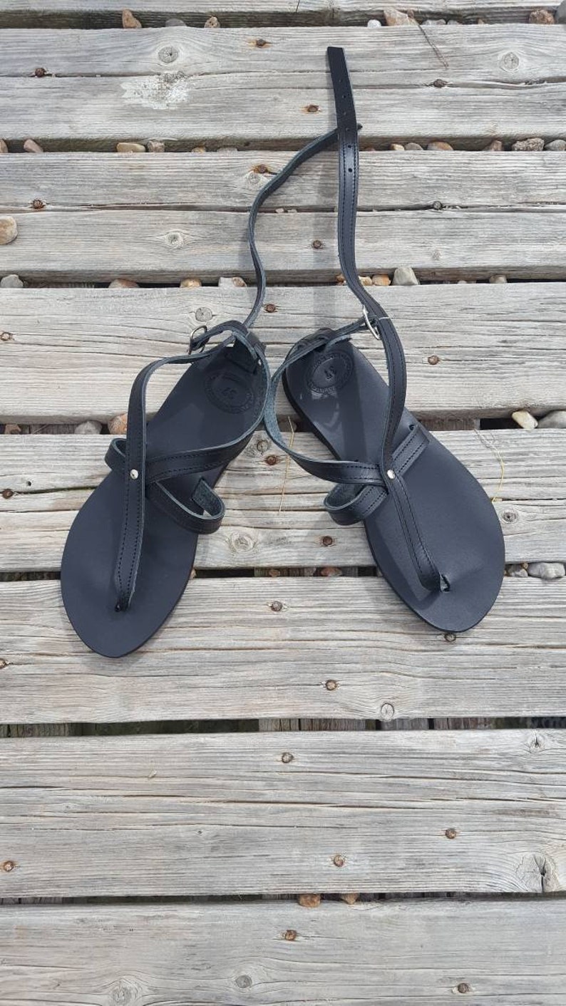 K&ouml;nnte beinhalten: Ein Paar schwarze Ledersandalen mit Riemen, die &uuml;ber Fu&szlig; und Kn&ouml;chel verlaufen. Die Sandalen befinden sich auf einer Holzoberfl&auml;che.