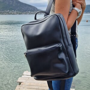 May include: A black leather backpack with a zippered front pocket. The backpack is being worn by a person standing on a wooden dock.