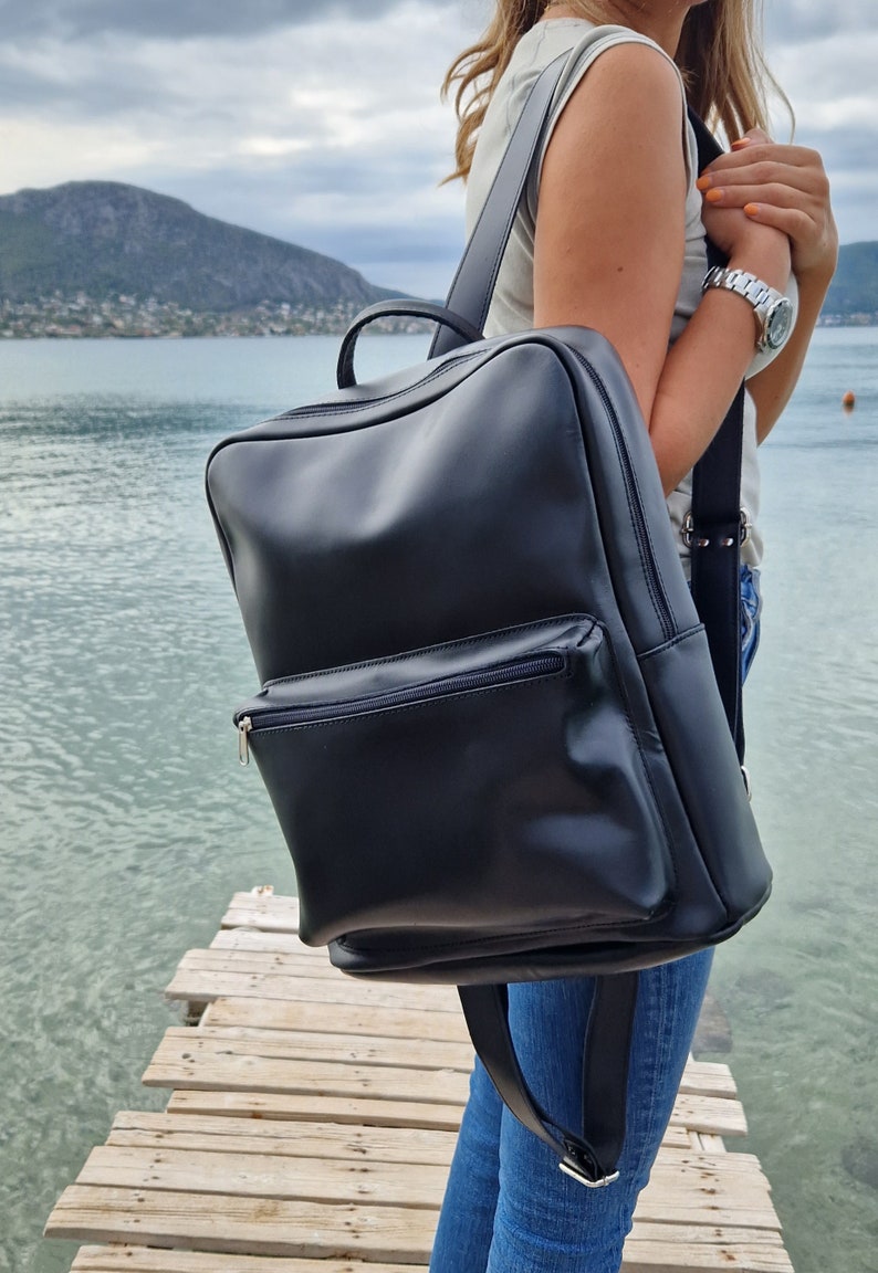 May include: A black leather backpack with a zippered front pocket. The backpack is being worn by a person standing on a wooden dock.