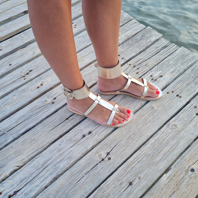 May include: A pair of gold leather sandals with ankle straps. The sandals have a simple design with a flat sole and a toe strap.