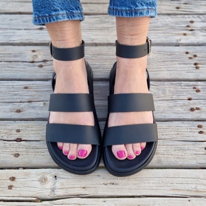 May include: A pair of black leather sandals with two straps across the foot. The sandals have a buckle closure at the ankle and a thick platform sole.