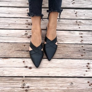 May include: A pair of black leather pointed-toe mules with criss-cross straps. The shoes are on a wooden surface.