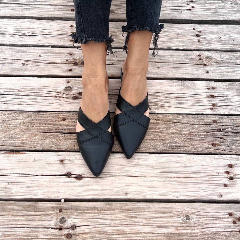 May include: A pair of black leather pointed-toe mules with criss-cross straps. The shoes are on a wooden surface.