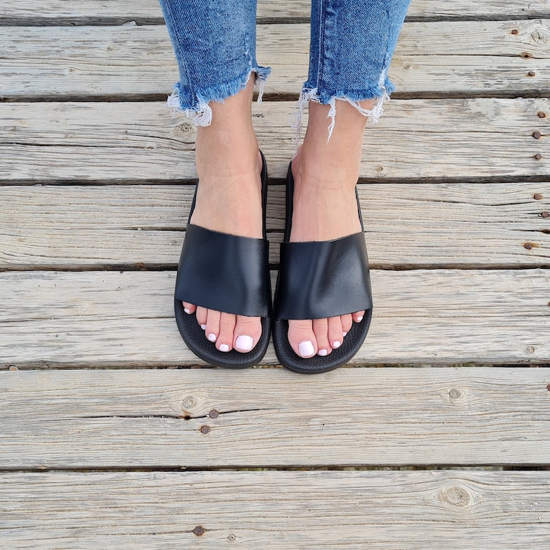 May include: A pair of black leather slide sandals with a simple design. The sandals are worn on a wooden surface.