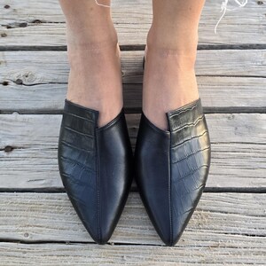 May include: A pair of black leather slip-on shoes with a pointed toe and a crocodile texture on the vamp. The shoes are on a wooden deck.