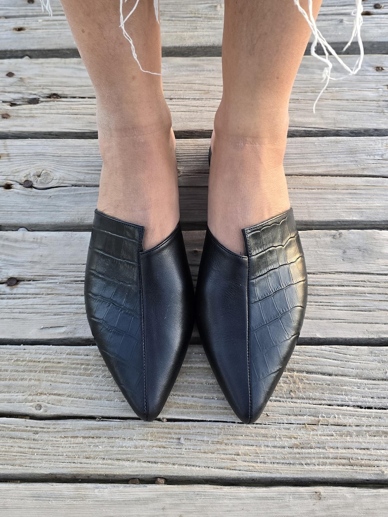 May include: A pair of black leather slip-on shoes with a pointed toe and a crocodile texture on the vamp. The shoes are on a wooden deck.
