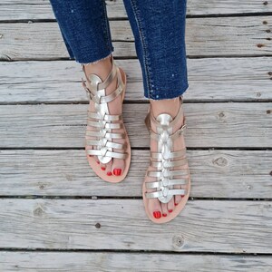 May include: A pair of gold gladiator sandals with a woven leather design. The sandals have a buckle closure and are worn on a wooden surface.