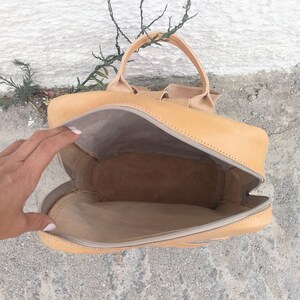 May include: A tan leather backpack with a zipper closure and a top handle. The backpack is open and the inside is visible.