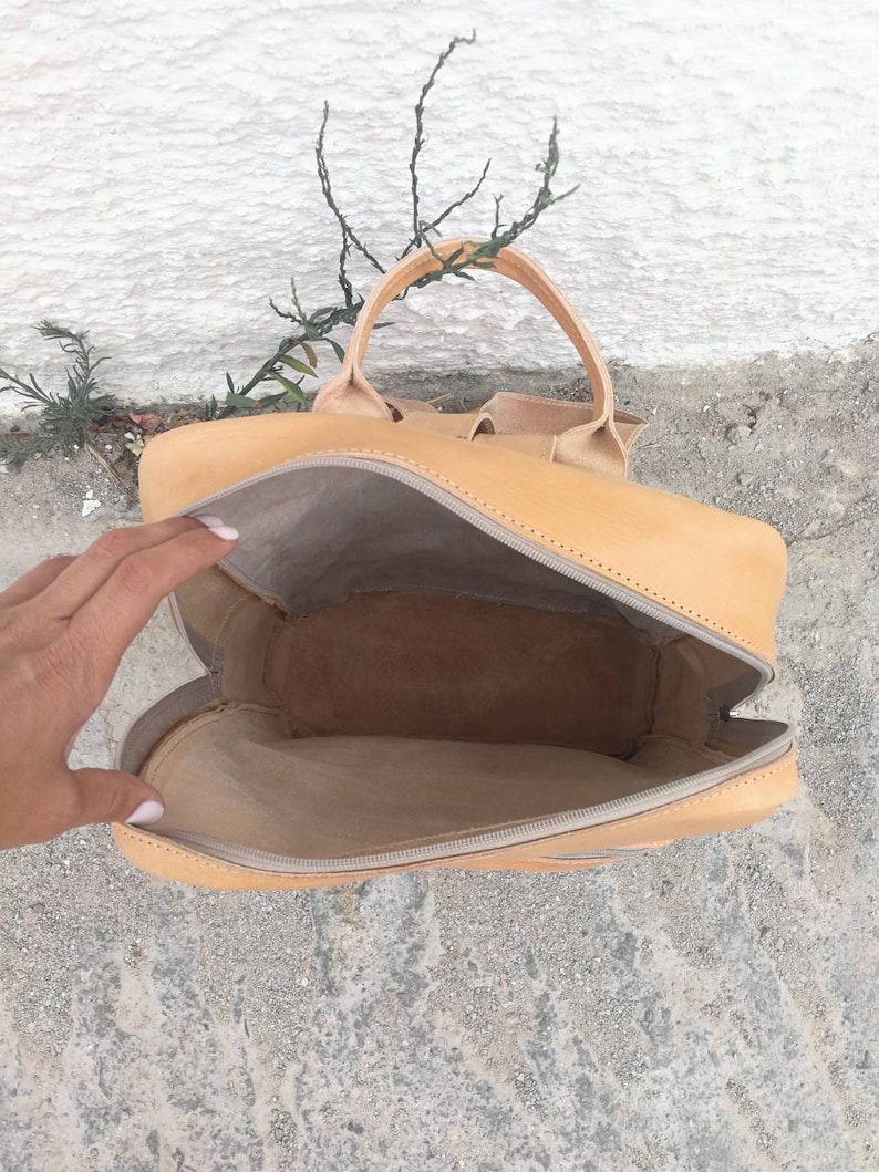 May include: A tan leather backpack with a zipper closure and a top handle. The backpack is open and the inside is visible.