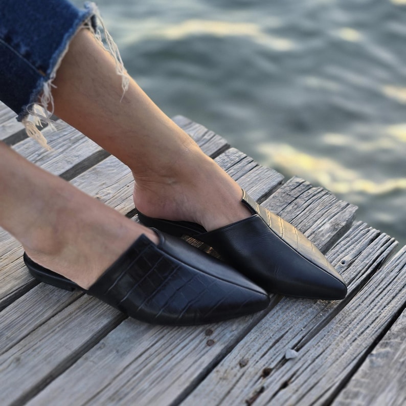 May include: Black leather slip-on mules with a pointed toe. One mule has a textured, crocodile-like pattern. The shoes are on a wooden surface, with a blurred water background.