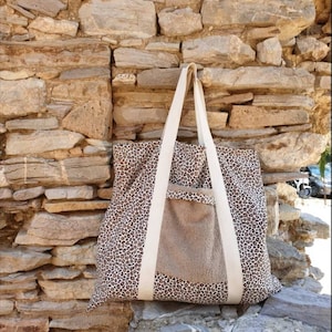 May include: A large tote bag with a brown and white leopard print design. The bag has a large pocket on the front with a brown fabric lining. The bag has white straps.