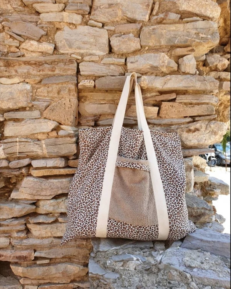 May include: A large tote bag with a brown and white leopard print design. The bag has a large pocket on the front with a brown fabric lining. The bag has white straps.