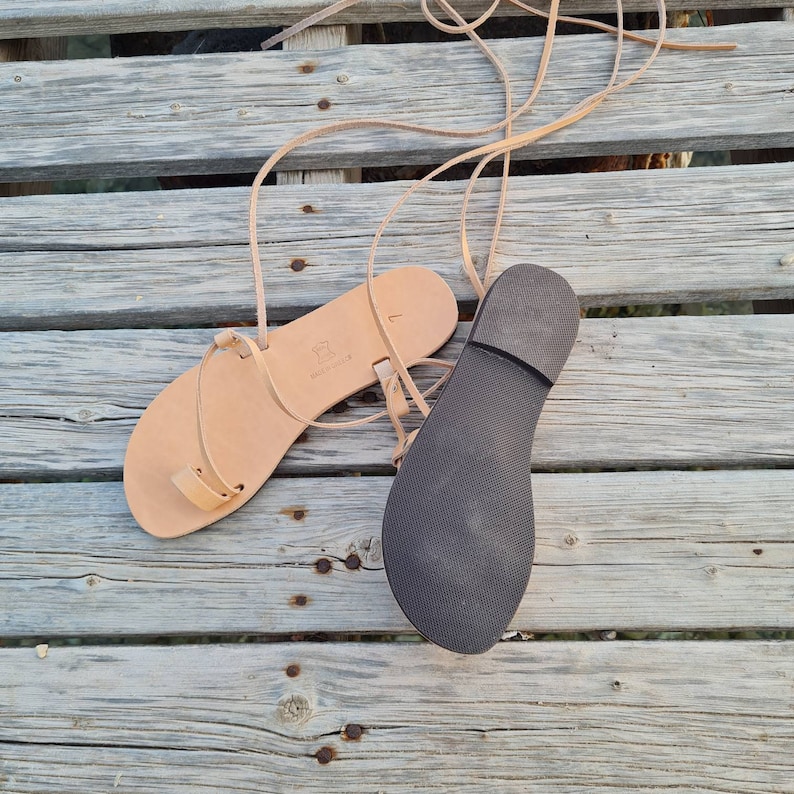 May include: A pair of beige leather sandals with long, thin straps. The sandals have a simple design with a toe loop and ankle straps. The soles are dark gray. The sandals are displayed on a weathered wooden surface.