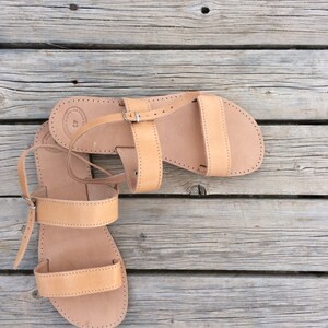 May include: A pair of tan leather sandals with a buckle closure. The sandals are on a wooden surface.