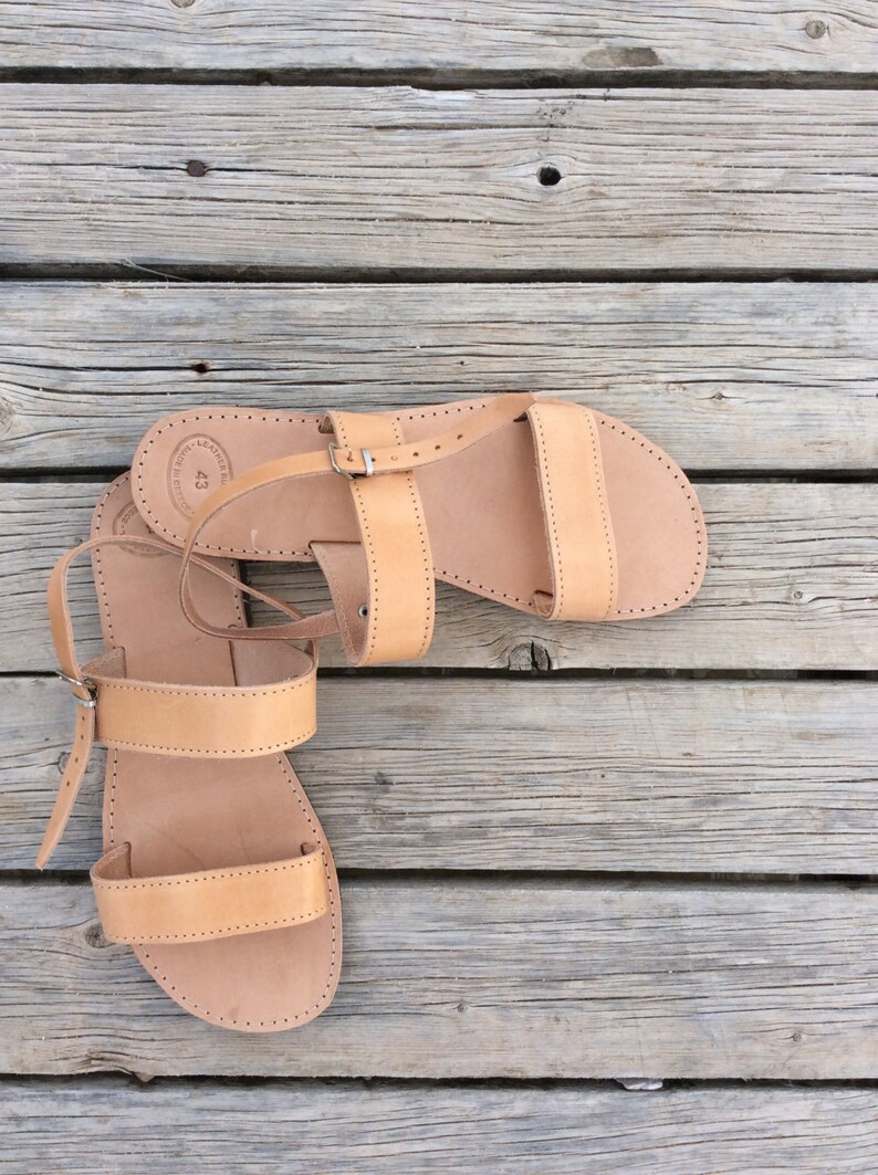 May include: A pair of tan leather sandals with a buckle closure. The sandals are on a wooden surface.