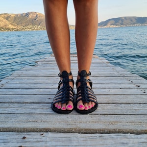 May include: A pair of black leather gladiator sandals with a criss-cross strap design. The sandals have a buckle closure and are worn on a wooden dock.