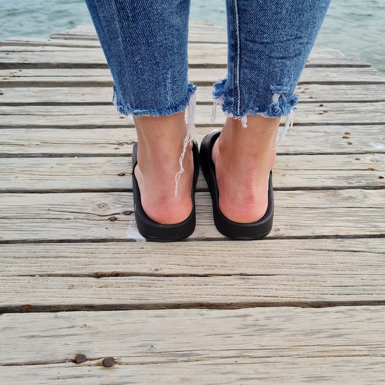 May include: A pair of black flip-flops with a person's feet wearing them. The person is wearing blue jeans with frayed hems.