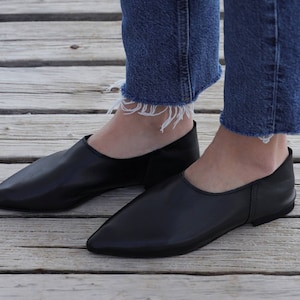 May include: A pair of black leather slip-on shoes with pointed toes. The shoes are worn by a person wearing blue jeans.