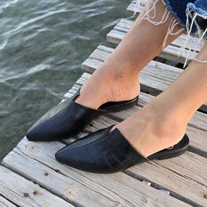 May include: Black leather slip-on shoes with pointed toes. One shoe has a textured pattern, while the other has a smooth surface. The shoes are on a wooden dock near water. The shoes are a size 8.