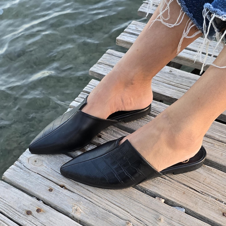 May include: Black leather slip-on shoes with pointed toes. One shoe has a textured pattern, while the other has a smooth surface. The shoes are on a wooden dock near water. The shoes are a size 8.