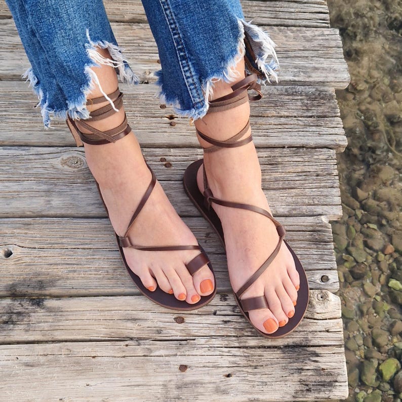 May include: Brown leather sandals with wrap-around ankle straps. The sandals have a toe loop design and are worn with distressed blue jeans. The image is taken outdoors on a wooden dock.