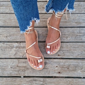 May include: Gold-colored strappy sandals with a toe loop and ankle ties. The sandals are worn with blue denim jeans with frayed edges. The sandals are on a wooden surface.