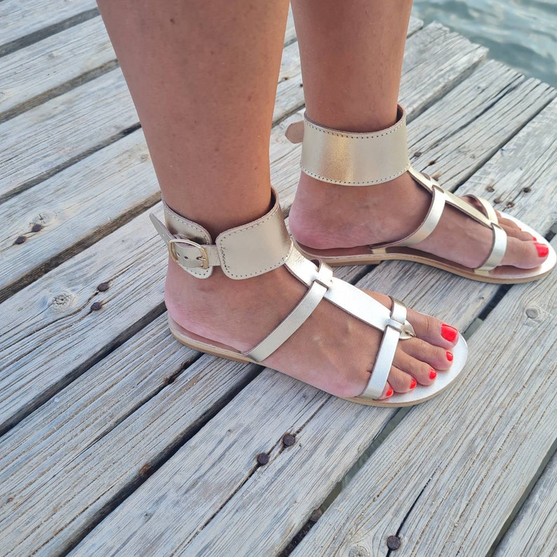 May include: Gold leather sandals with ankle straps and buckle closures. The sandals have a flat sole and are worn on a person's feet.
