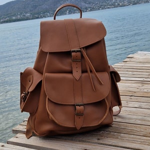 Puede incluir: Una mochila de cuero marr&oacute;n con varios bolsillos y hebillas. La mochila est&aacute; sobre una superficie de madera.