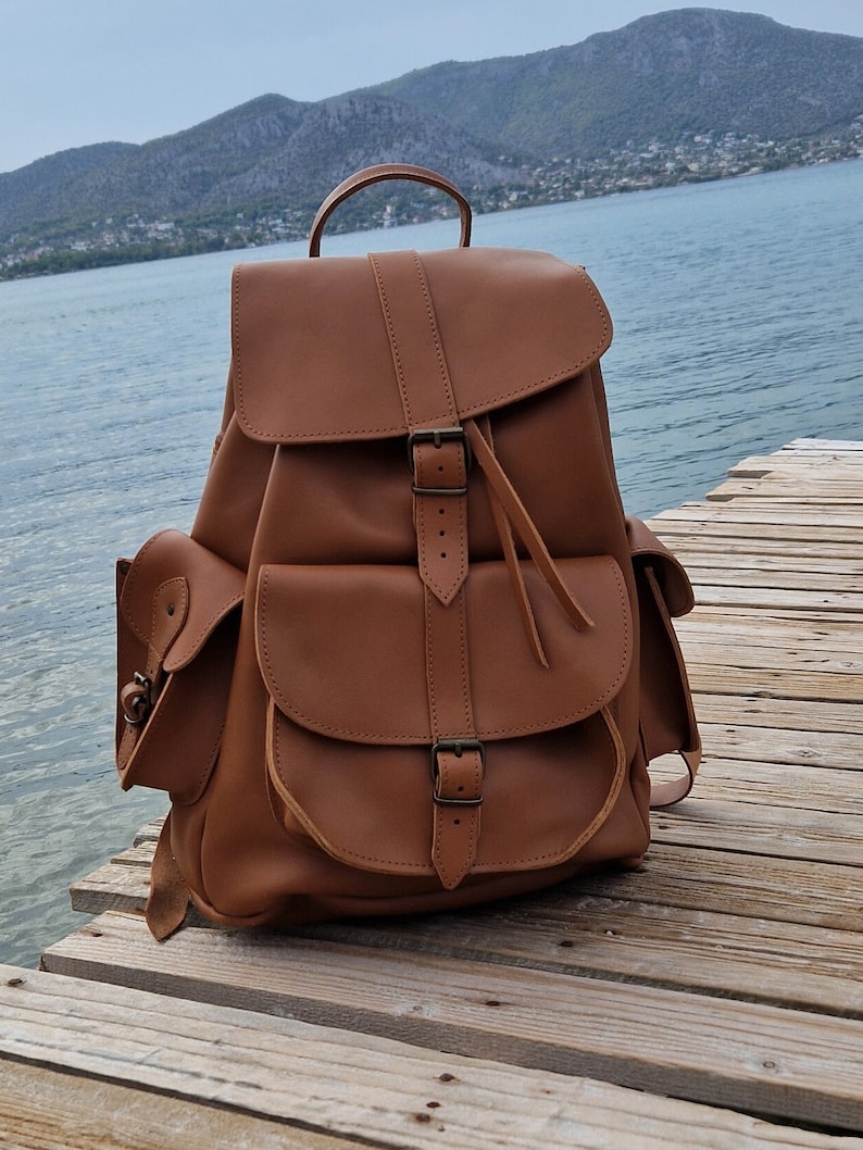 Puede incluir: Una mochila de cuero marr&oacute;n con varios bolsillos y hebillas. La mochila est&aacute; sobre una superficie de madera.
