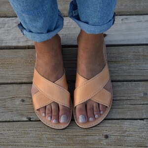 May include: A pair of light brown leather sandals with criss-cross straps. The sandals are worn by a person with bare feet and are on a wooden surface.