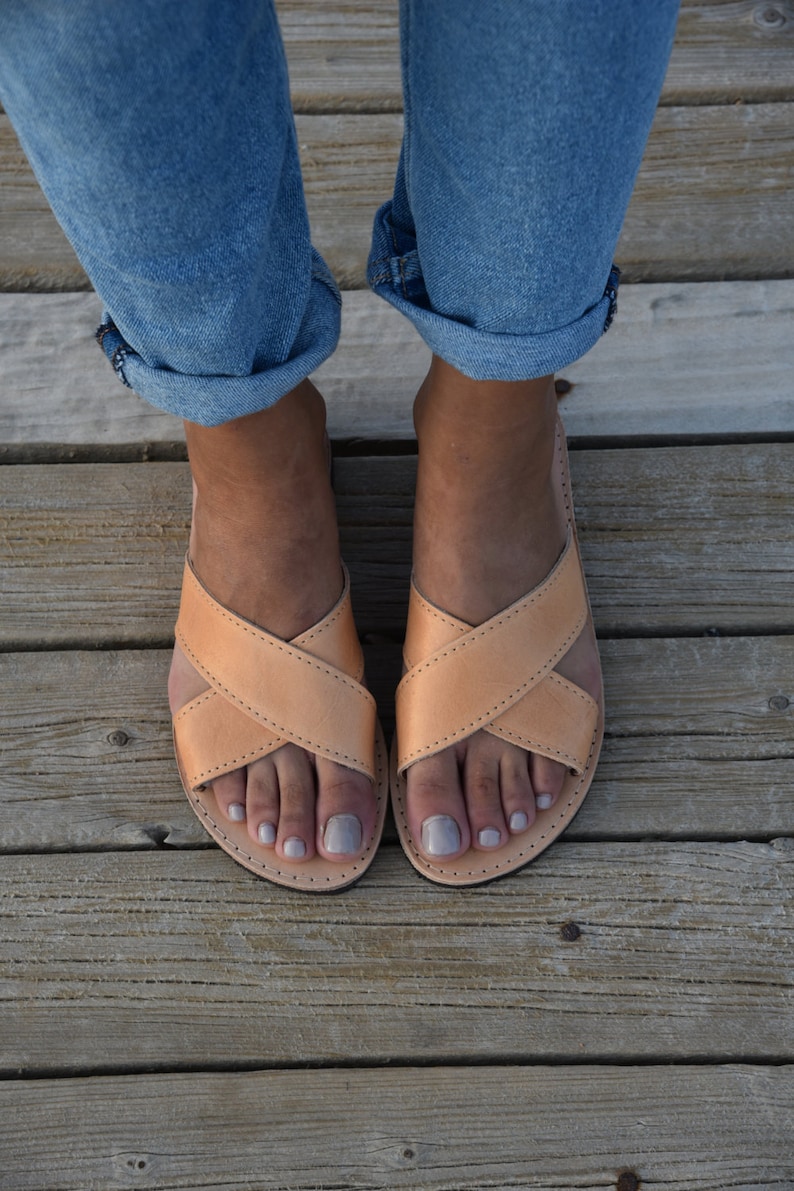 May include: A pair of light brown leather sandals with criss-cross straps. The sandals are worn by a person with bare feet and are on a wooden surface.