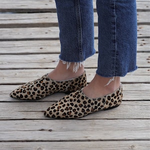 May include: A pair of brown and black leopard print flats with pointed toes. The shoes are worn by a person wearing blue jeans.
