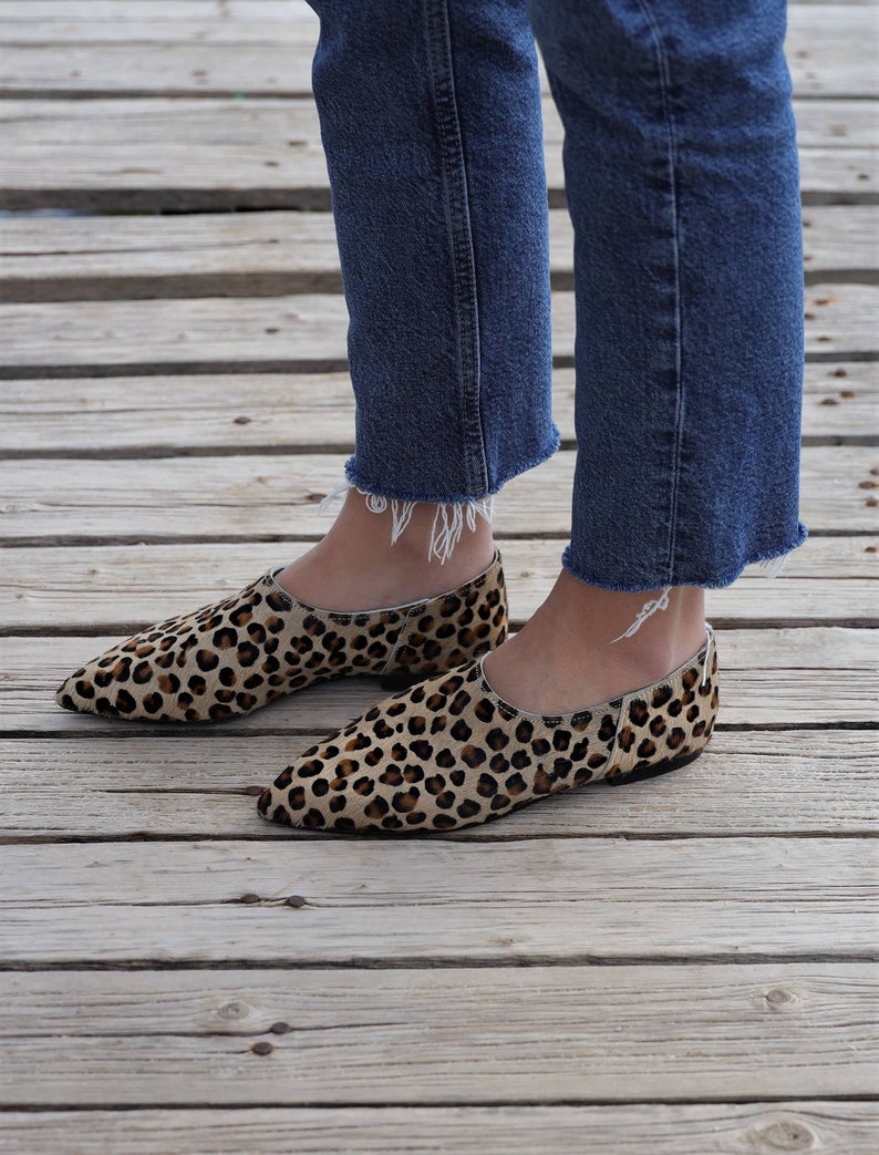 May include: A pair of brown and black leopard print flats with pointed toes. The shoes are worn by a person wearing blue jeans.