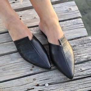 May include: A pair of black leather mules with a pointed toe and a crocodile-textured vamp. The shoes are on a wooden surface, showcasing their sleek design and style. The open-back design offers a modern, slip-on aesthetic.