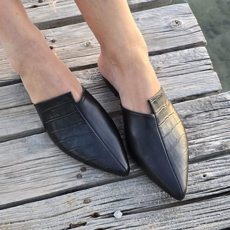 May include: A pair of black leather mules with a pointed toe and a crocodile-textured vamp. The shoes are on a wooden surface, showcasing their sleek design and style. The open-back design offers a modern, slip-on aesthetic.