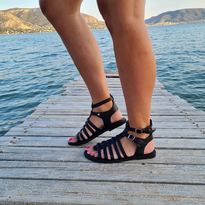 May include: Black leather gladiator sandals with multiple straps and buckles. The sandals are worn by a person standing on a wooden dock.