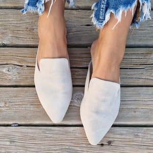 May include: A pair of light beige suede slip-on mules with pointed toes. The shoes are worn by a person with bare feet and blue jeans.