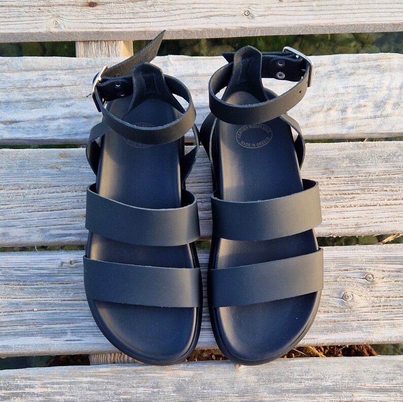 May include: A pair of black leather sandals with two straps across the foot and an ankle strap. The sandals have a simple design and are made in Greece.