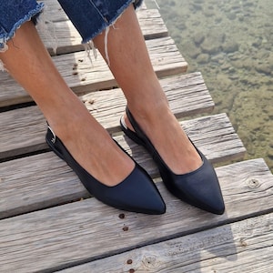 May include: A pair of black leather slingback flats with pointed toes. The shoes are worn by a person standing on a wooden dock.